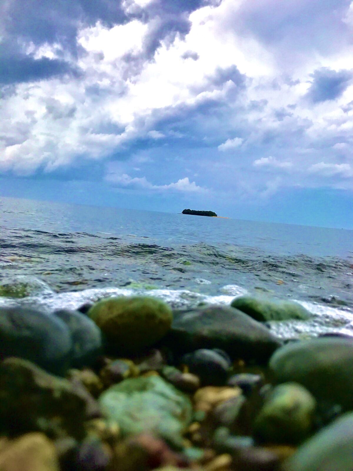 This photo captures a stunning coastal scene, where nature's contrasts create a dramatic and serene atmosphere. In the foreground, a rugged shoreline is adorned with an array of rocks—some smooth, some jagged—partially submerged as waves gently lap against them. The distant island serves as a focal point, standing quietly amid the vast ocean, its silhouette adding a sense of mystery and solitude. Above, the sky is alive with thick, dynamic clouds, layering light and shadow to create depth and movement. The interplay between the moody sky and the tranquil sea evokes a powerful sense of resilience and balance—a theme you seem to appreciate in creative expression. This image feels like a reflection of harmony in nature, where stillness and motion coexist beautifully. This photo is located in Maangas Presentacion Camarines Sur, Philippines.