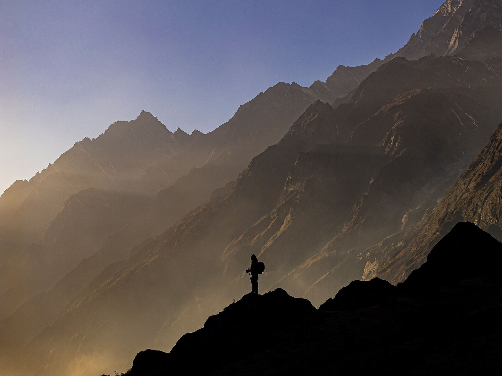 In the heart of Nepal’s Langtang National Park, a solitary tourist stands silhouetted against the fading light of dusk, surrounded by the majestic layers of the Himalayas. The photograph captures the surreal beauty of sunset as soft rays filter through the mountain ridges, painting the valley in hues of amber and blue. The image evokes a sense of awe, reflection, and quiet resilience in the face of nature’s vastness.