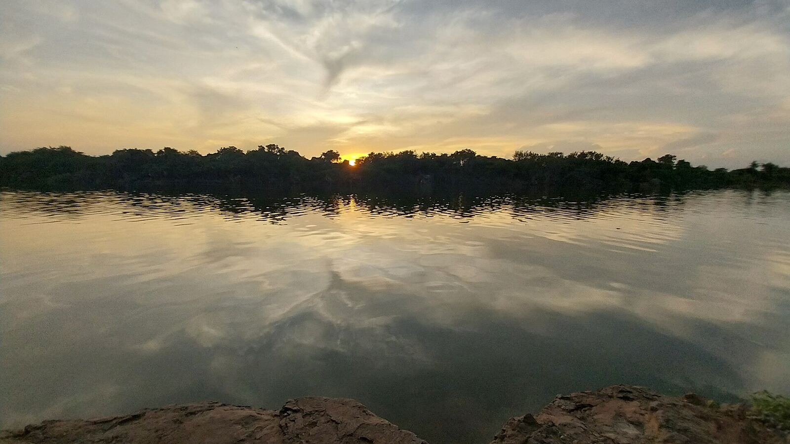 This photograph captures a tranquil sunset scene along the Godavari River near Raorajur, Maharashtra (431514). The setting sun is partially hidden behind a dense line of trees along the riverbank, casting warm golden and amber hues across the sky and water. The soft, diffused light suggests it was taken during the late evening, just before dusk, when the atmosphere is calm and serene. The surface of the river is gently rippled, creating shimmering reflections of the sky, trees, and fading sunlight. These subtle waves add texture to the water while still maintaining an overall sense of stillness. In the foreground, natural rocky formations along the riverbank are visible, grounding the composition and providing contrast to the smooth water. The background consists of thick greenery and forested land along the horizon, highlighting the natural and untouched environment of the location. The overall mood of the image is peaceful, reflective, and meditative, evoking a sense of solitude and connection with nature.